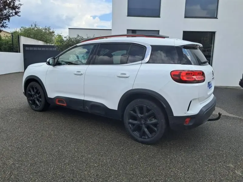 Vue côté gauche arrière d'un Citroën C5 Aircross blanc 2019 avec jantes noires et inserts orange sur bas de caisse.