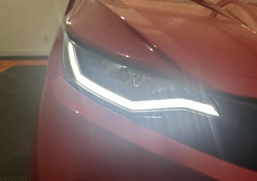 Gros plan sur le phare avant gauche allumé d’une Aixam Ambition 0.5 City Sport rouge, année 2026.