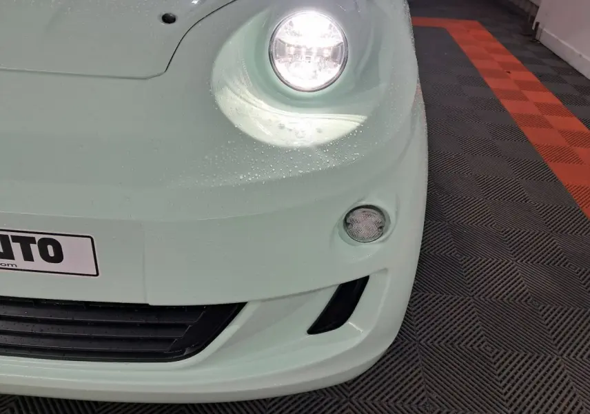 Gros plan sur l'avant droit vert clair de l'AIXAM MINAUTO E chic 2026 avec phare allumé et gouttes d'eau visibles.