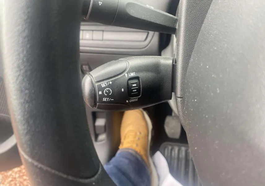 Gros plan sur la commande de régulateur de vitesse au volant d'une Peugeot 208 gris shark, avec pédale et chaussure visible.