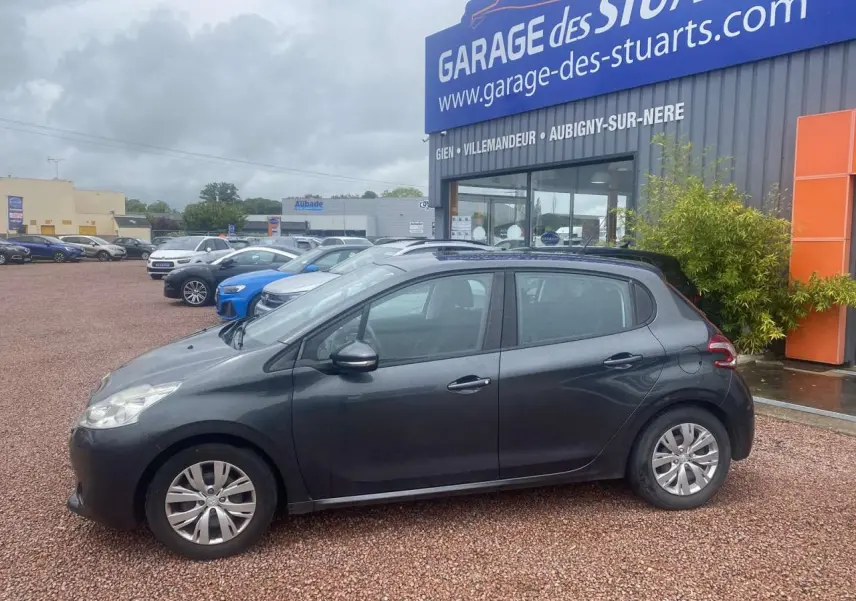 Profil côté gauche d'une Peugeot 208 gris shark 2013 garée devant un garage avec jantes acier et poignées noires.