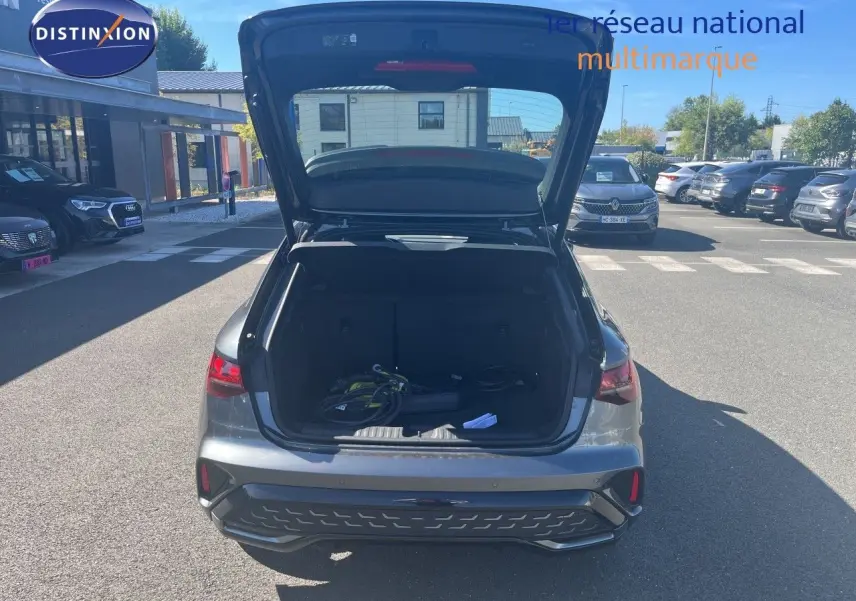Vue arrière d'une Audi A3 Sportback gris Daytona avec coffre ouvert montrant câbles de recharge dans un parking extérieur.