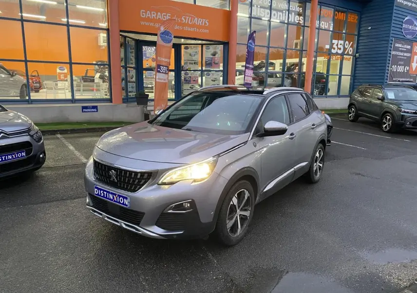 Peugeot 3008 gris Artense vue 3/4 avant droit avec phare allumé, stationnée devant un garage commercial.