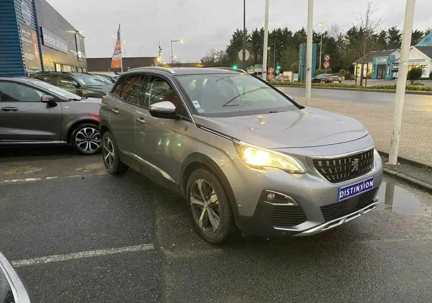 Peugeot 3008 gris Artense vue 3/4 avant droit avec phares allumés sur parking urbain.