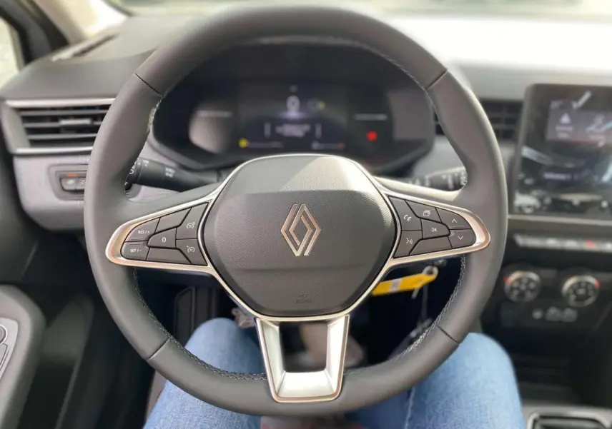 Vue rapprochée du volant multifonction noir de la Renault Clio 2025 avec tableau de bord et écran tactile en arrière-plan.