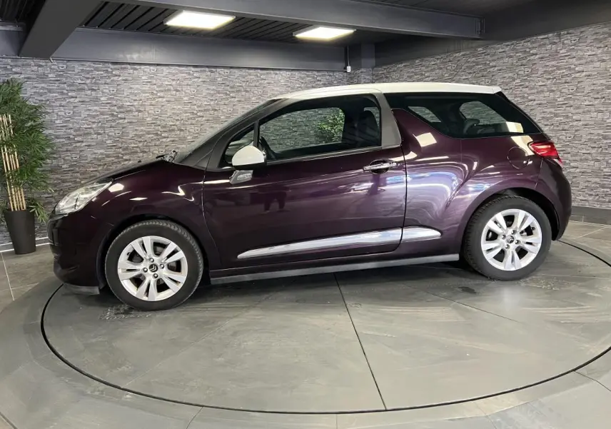 Profil côté gauche d'une DS3 violet avec toit blanc, jantes alliage et poignées chromées en intérieur showroom.