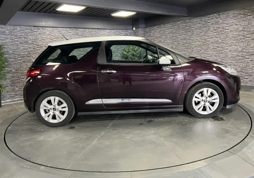 Profil côté gauche d'une DS3 violet avec toit blanc et jantes alliage dans un showroom intérieur.