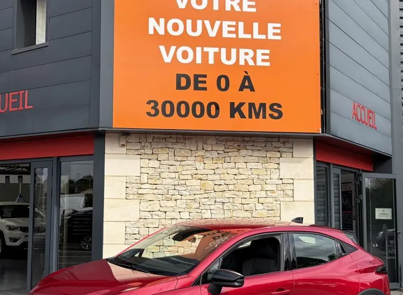 Renault Clio Evolution rouge vue en 3/4 avant droit, avec jantes noires et calandre distinctive, devant un bâtiment commercial.