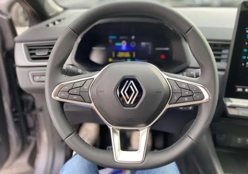 Vue rapprochée du volant multifonction noir avec logo Renault, tableau de bord numérique flou en arrière-plan.