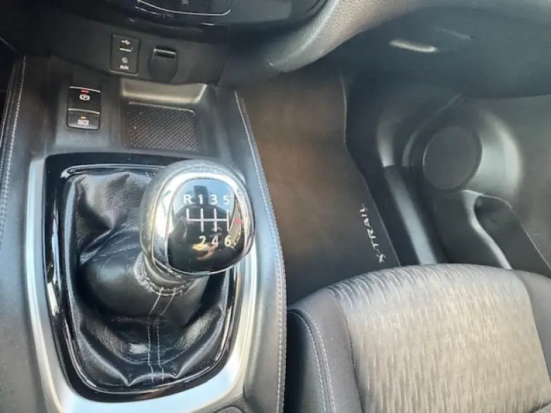 Gros plan sur le levier de vitesses manuel du Nissan X-TRAIL noir, avec tapis de sol marqué et siège tissu gris visible.