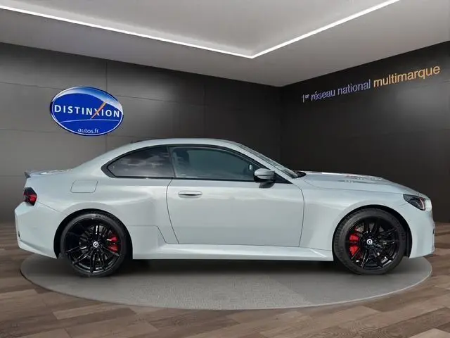 Profil droit d'une BMW M2 G87 gris Brooklyn Grau avec jantes noires et étriers de frein rouges dans un showroom.