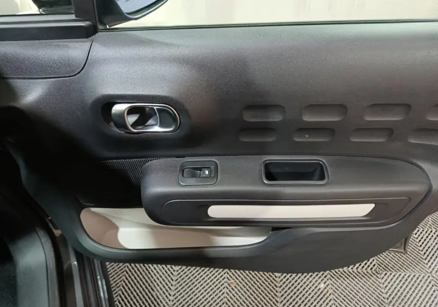 Vue intérieure côté gauche de la porte d'une Citroën C3 2024 gris foncé, avec poignée chromée et commandes électriques.