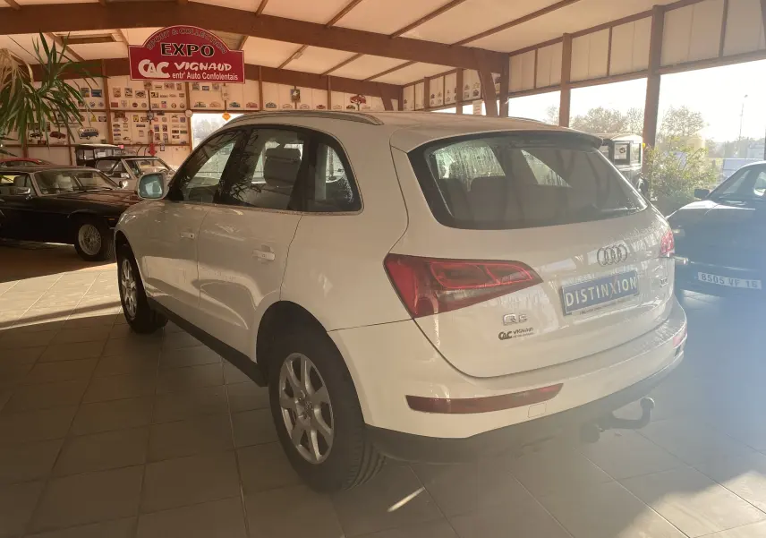Audi Q5 blanc en 3/4 arrière droit dans un showroom, avec attelage visible et plaques d'exposition.