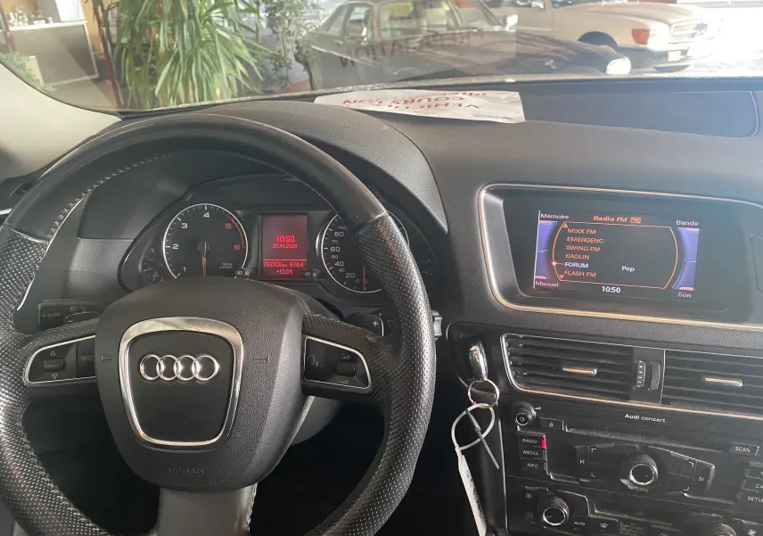 Intérieur de l’Audi Q5 blanc 2012, vue du poste de conduite avec volant en cuir et écran multimédia allumé.