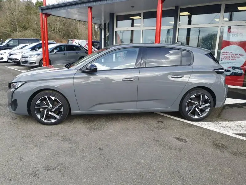 Profil côté gauche d'une Peugeot 308 gris Sélénium 2024 avec jantes alliage et vitres surteintées, stationnée devant un bâtiment.