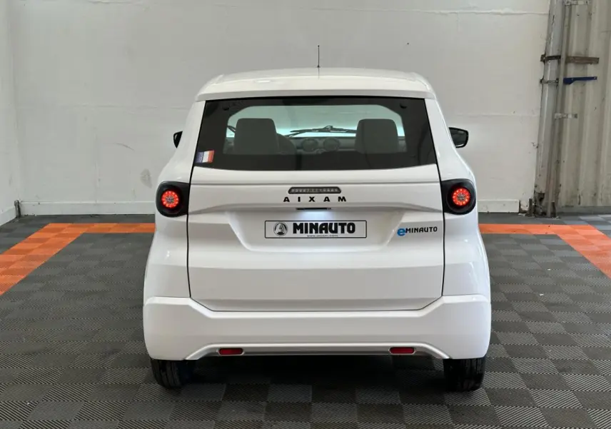 Vue arrière d'une AIXAM MINAUTO E ACCESS blanche 2026 avec feux ronds et logo MINAUTO visible sur un sol à damier noir et orange.