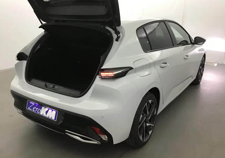 Vue 3/4 arrière droite d'une Peugeot 308 blanche Okenite avec coffre ouvert et feux arrière allumés dans un studio.