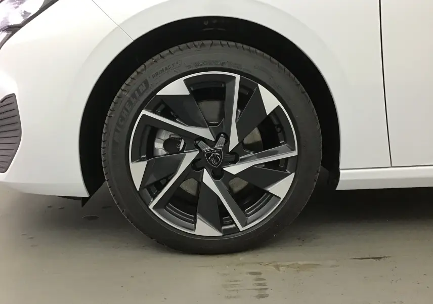 Gros plan sur la roue avant gauche d'une Peugeot 308 blanche Okenite avec jante alliage noire et argentée au design moderne.