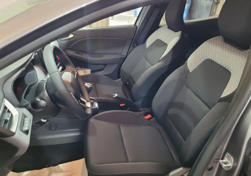 Vue intérieure côté conducteur de la Renault Clio gris foncé 2025, sièges tissu bicolore et volant multifonctions cuir synthétique.