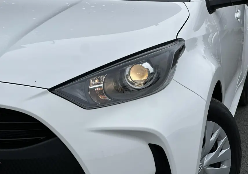 Gros plan sur le phare avant droit d'une Toyota Yaris blanche 2025, soulignant la calandre et la jante acier.