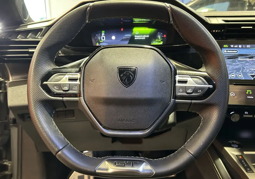 Volant cuir perforé noir avec logo Peugeot et badge GT, tableau de bord numérique et écran GPS visibles en arrière-plan.