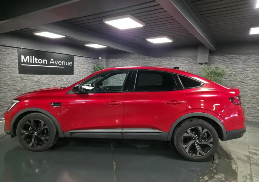 Profil droit du Renault Arkana 2023 rouge flamme avec jantes noires et toit ouvrant électrique visible en intérieur showroom.