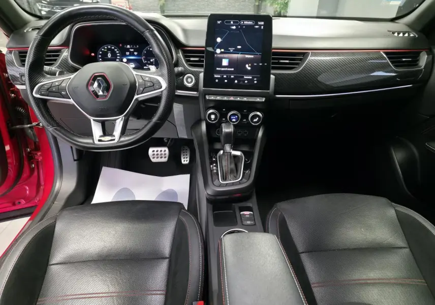 Vue intérieure avant du Renault Arkana rouge, tableau de bord avec écran tactile vertical et volant sport cuir noir à surpiqûres rouges.