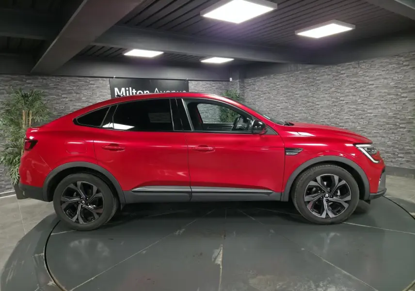Profil droit d'une Renault Arkana rouge flamme 2023 avec jantes noires et toit ouvrant électrique visible en intérieur showroom.