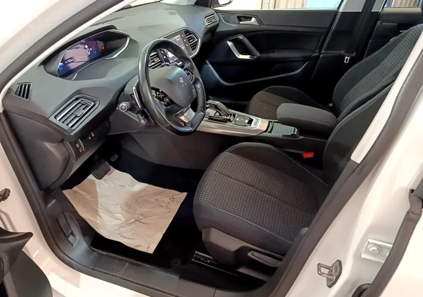 Intérieur avant droit de la Peugeot 308 blanche, montrant le volant cuir, console centrale et sièges noirs tissu.