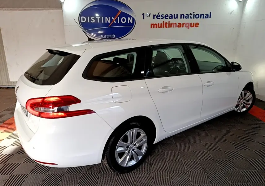 Peugeot 308 break blanc vue 3/4 arrière droit, feux arrière allumés, stationné en intérieur sur sol à motifs.