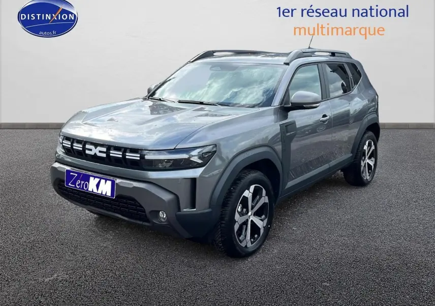 Dacia Duster gris schiste en 3/4 avant droit avec jantes alu et barres de toit visibles sur fond neutre.
