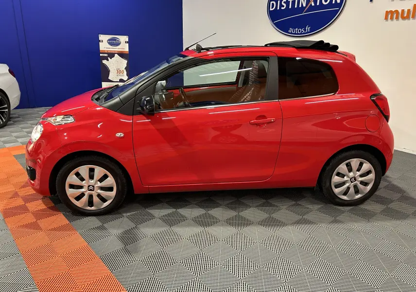 Vue de profil côté gauche d'une Citroën C1 Airscape rouge avec toit ouvrant en toile replié, dans un showroom.