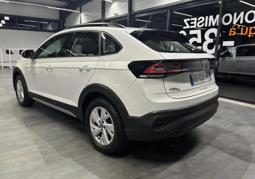 Vue 3/4 arrière droite d’un Volkswagen Taigo blanc avec jantes alliage et protections noires au bas de caisse en intérieur showroom.