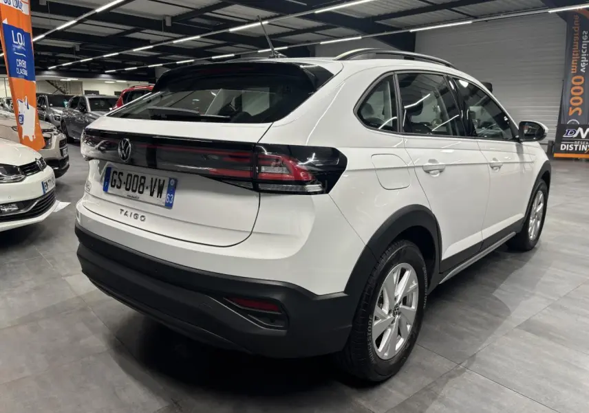 Vue 3/4 arrière droite d’un Volkswagen Taigo blanc avec feux arrière noirs et jantes alliage visibles en intérieur showroom.