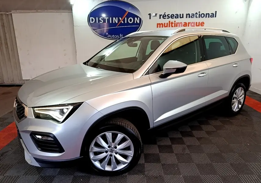 Vue 3/4 avant droit d’un SEAT Ateca gris clair 2022, avec jantes alliage et barres de toit chromées, en intérieur showroom.