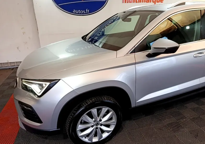 Vue de profil avant droit d’un SEAT Ateca gris clair avec jante alliage et feux LED allumés en intérieur showroom.