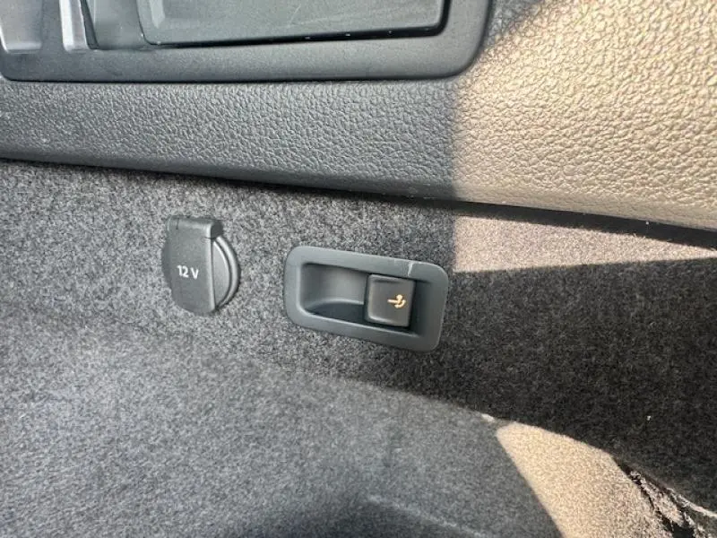Détail de la prise 12V et du bouton d'ouverture de banquette arrière dans le coffre d'une Volkswagen Passat SW gris manganèse métallisé.