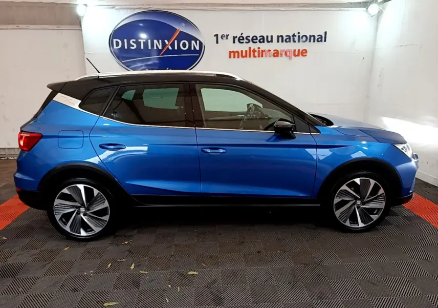 Vue latérale droite d'un SEAT Arona bleu 2022 avec toit blanc et jantes alliage distinctives en intérieur showroom.