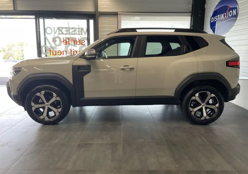 Profil côté gauche du SUV Dacia Duster beige Sandstone 2025 avec jantes alliage 18 pouces et barres de toit noires.