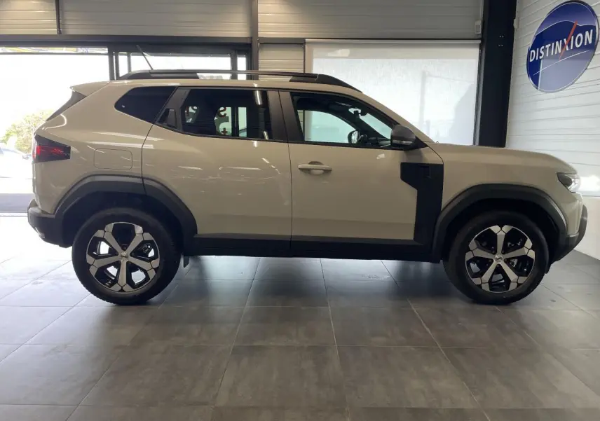 Profil droit du SUV Dacia Duster beige avec jantes alliage et barres de toit noires en intérieur showroom.