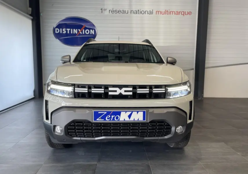 Vue frontale du SUV Dacia Duster beige Sandstone 2025 avec calandre noire et feux LED allumés en intérieur showroom.