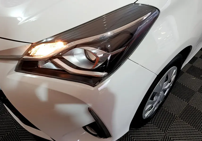 Gros plan sur le phare avant gauche allumé d'une Toyota Yaris blanche, avec partie de la roue visible.
