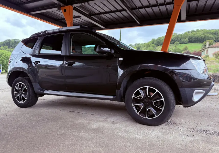 Vue de profil côté gauche d'un Dacia Duster noir métal 2017 sous abri avec jantes bi-ton et barres de toit.