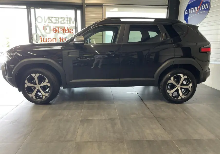 Profil côté gauche du Dacia Duster 2025 noir métallisé avec jantes alliage et barres de toit noires visibles en intérieur showroom.