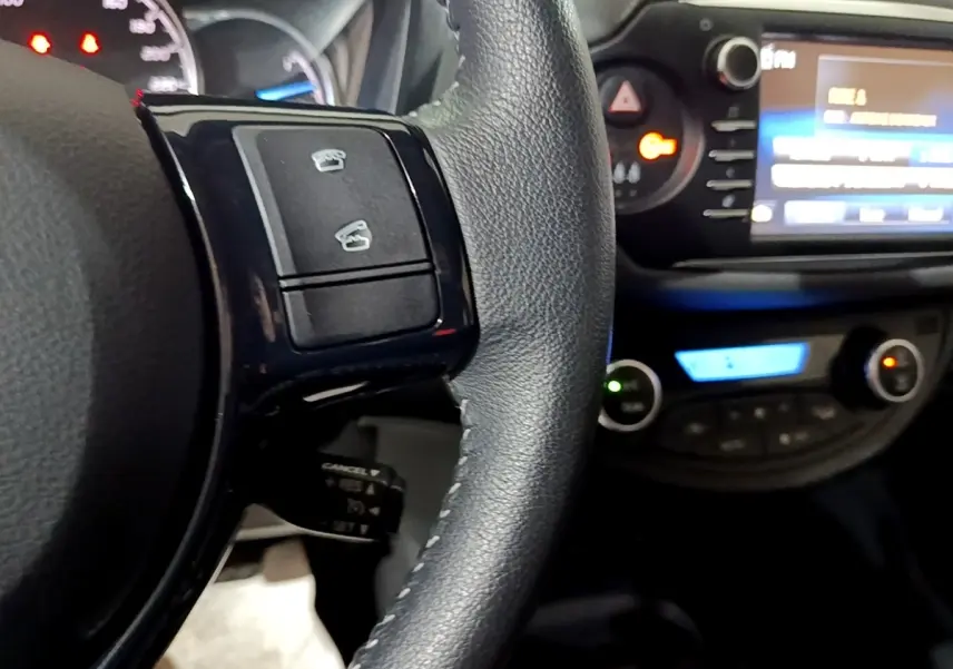 Gros plan sur le volant cuir et alu de la Toyota Yaris blanche, avec commandes multifonctions et tableau de bord éclairé.