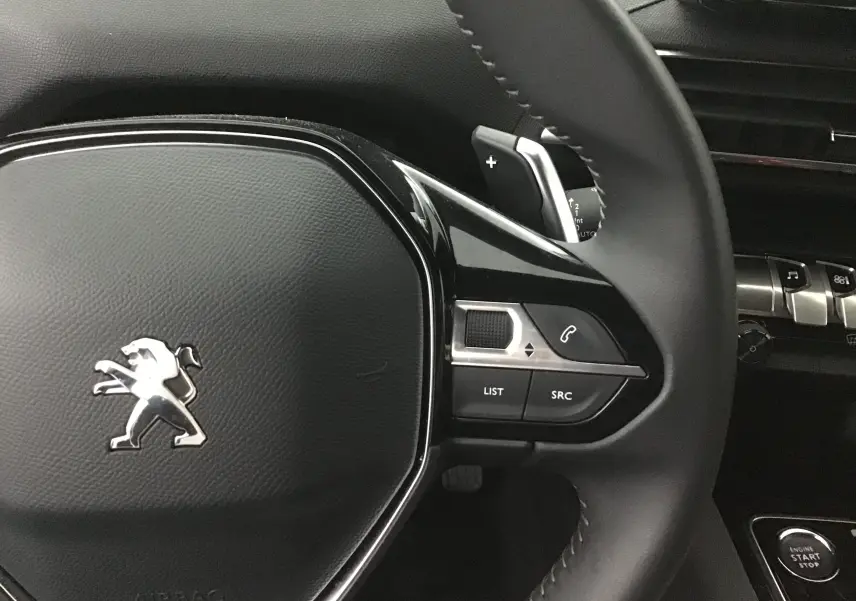Gros plan sur le volant noir du Peugeot 5008 2024 avec commandes intégrées et logo lion chromé au centre.
