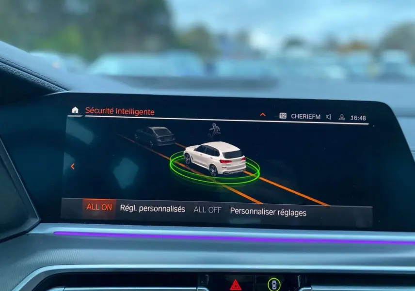 Écran intérieur du BMW X5 blanc 2022 montrant l'interface de sécurité intelligente avec visualisation 3D du véhicule en conduite.