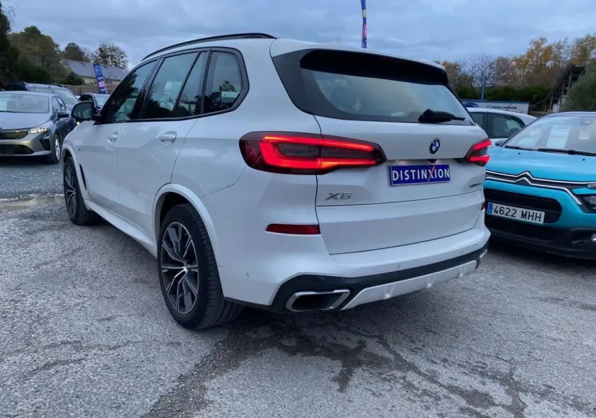 BMW X5 blanc vue 3/4 arrière droit avec feux allumés et double sortie d'échappement intégrée au pare-chocs.