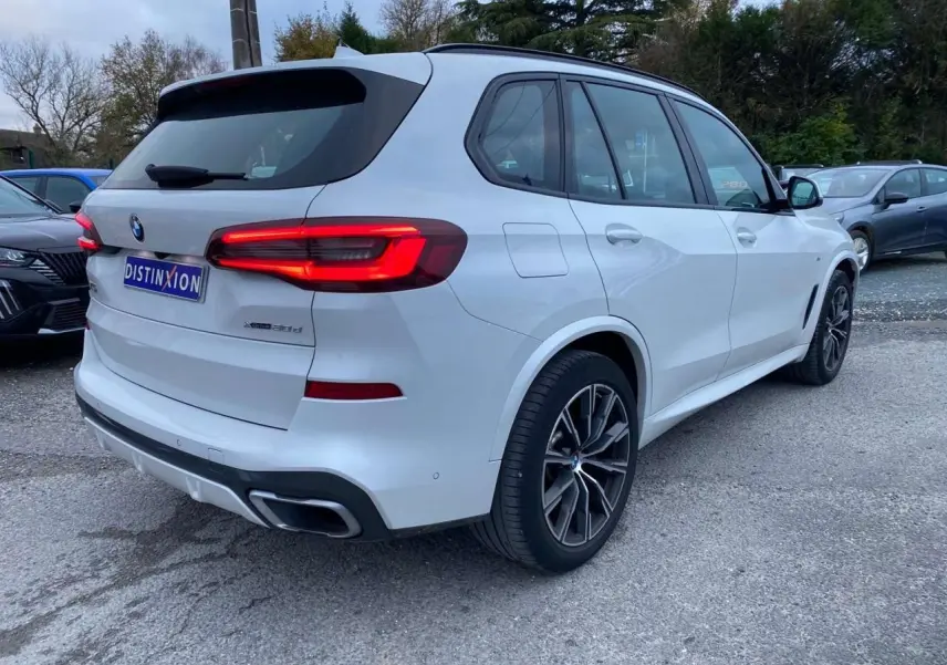 BMW X5 xDrive 30d blanc vu en 3/4 arrière droit avec jantes M Sport et feux arrière allumés.