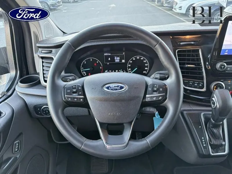 Vue intérieure centrée sur le volant cuir noir du Ford Transit Custom 2023 avec tableau de bord et levier de vitesses automatique.